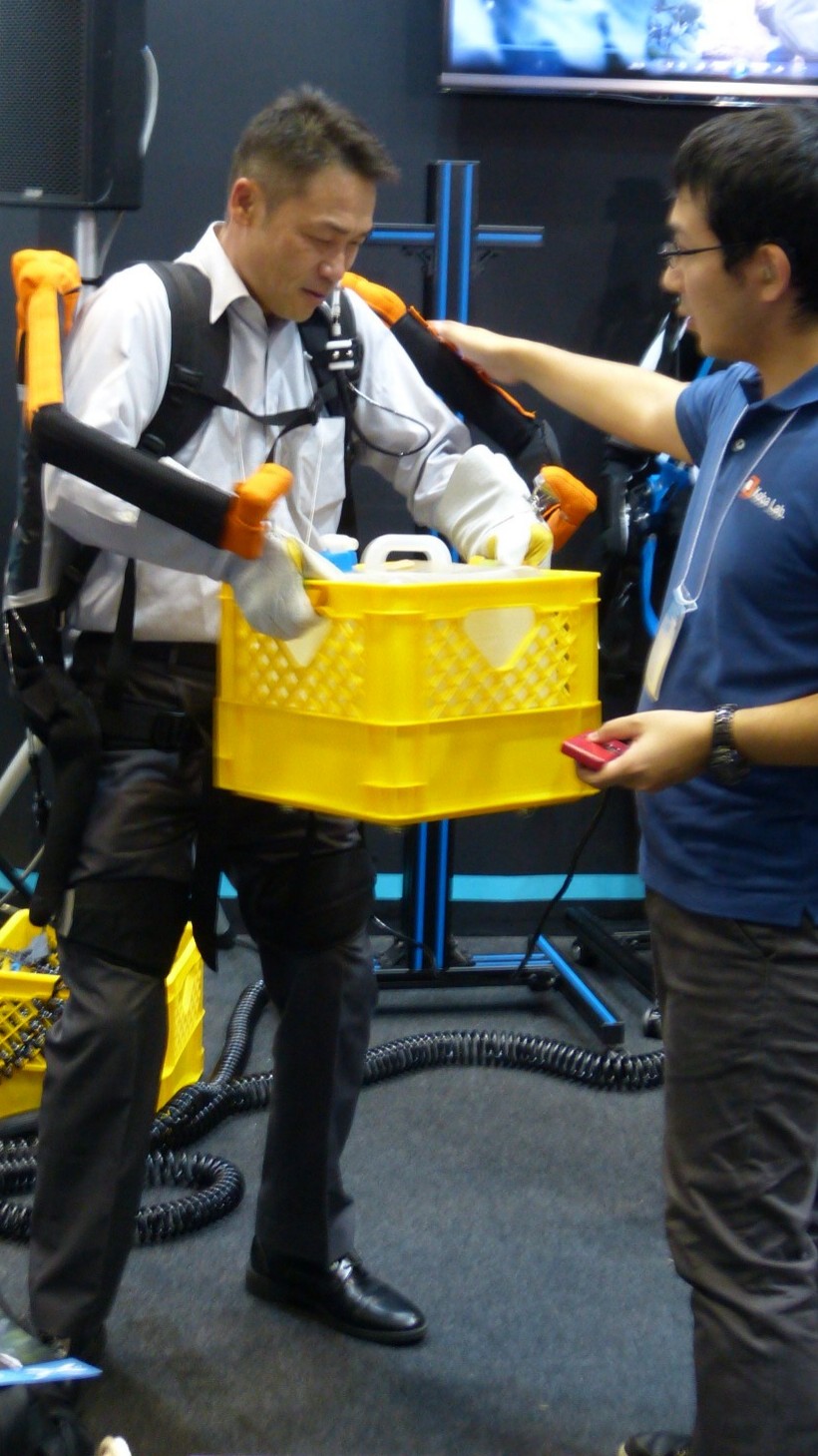 a man lifts a yellow plastic bin with assistance from an exoskeleton worn like a backpack